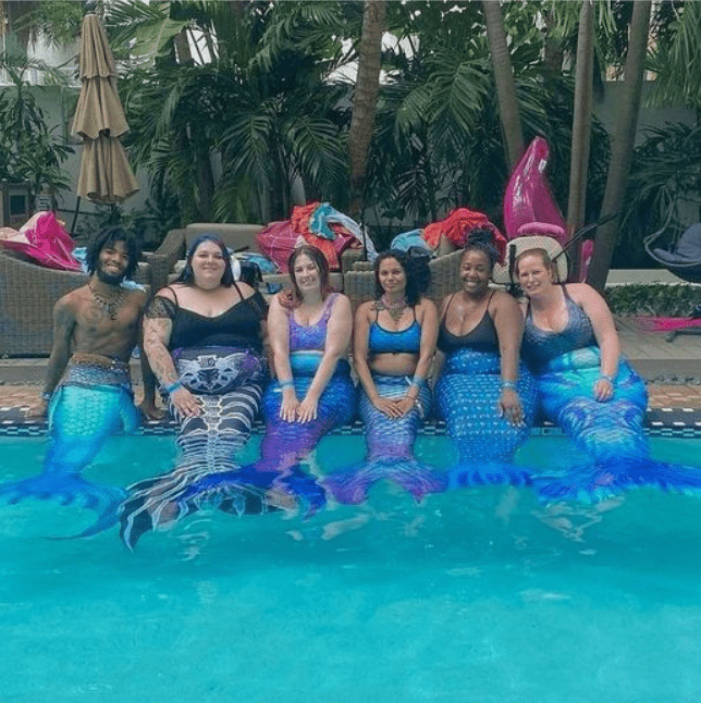 Various Black, African-American, and white merfolk in a pool with green trees in the background